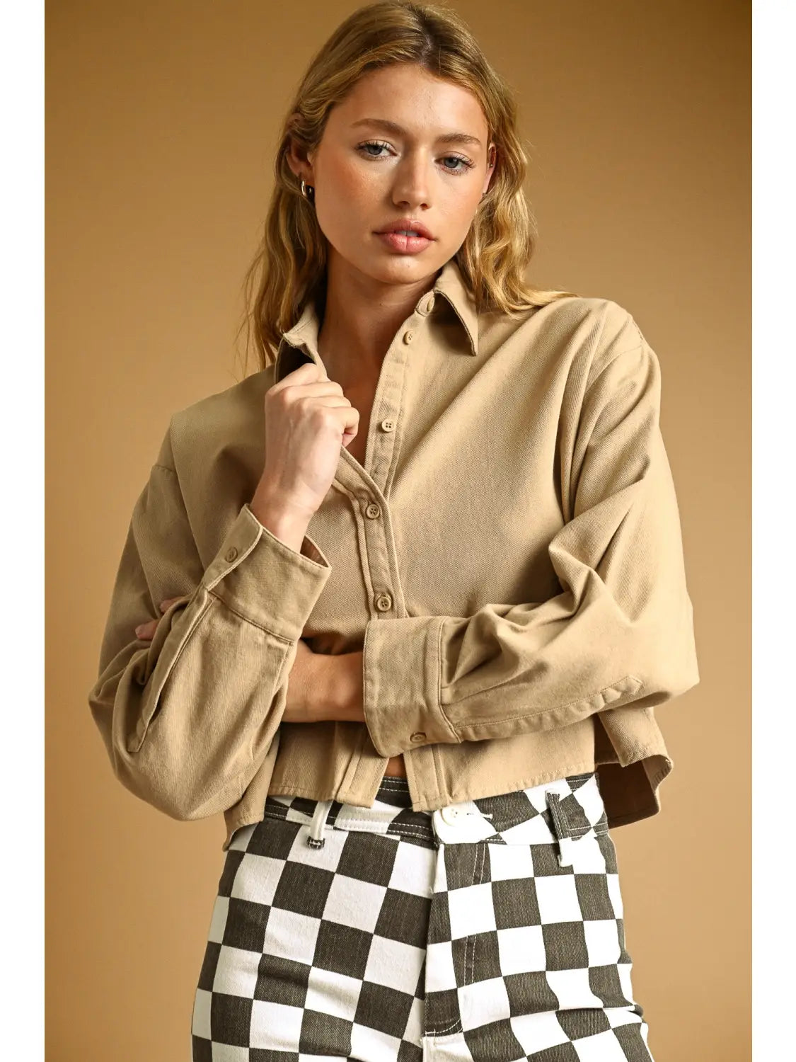 A woman modeling a cropped, long-sleeve button-up shirt in a warm tan/khaki color, paired with black and white checkered pants, against a solid warm brown studio background.