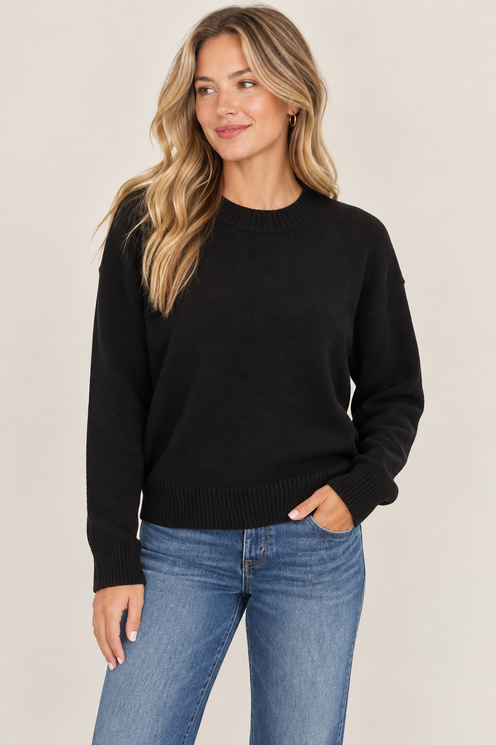 A woman wearing a black crewneck knit sweater and blue jeans, standing against a plain beige studio background.